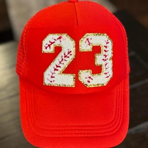 May include: Red trucker hat with a white and gold baseball number 23 with red stitching. The number is on a white background with a baseball stitch design.