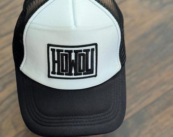 May include: A black and white trucker hat with a curved brim. The front panel is white with the word "HOWDU" in a black, blocky font. The mesh back is black, and the hat has a black button on top.
