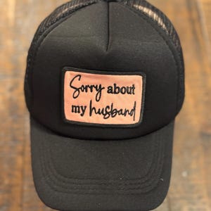 May include: Black trucker hat with a mesh back and a curved brim. The hat features a rectangular patch with the text "Sorry about my husband" in a cursive font. The patch is a light pink color.