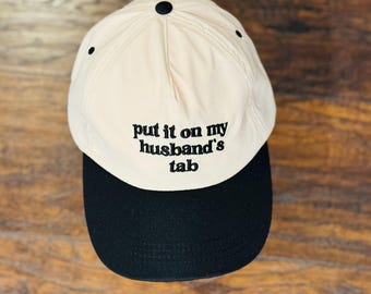May include: A beige baseball cap with a black brim. The cap features the embroidered phrase "put it on my husband's tab" in black lettering. The cap has a curved brim and a button on top.