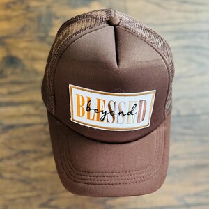 May include: Brown trucker hat with a mesh back and a curved brim. The front panel features a rectangular patch with the word "BLESSED" in orange and white, with "beyond" in a script font.