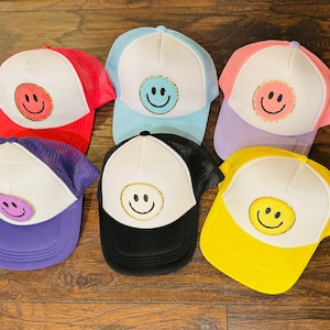 May include: Six trucker hats with different colored brims and mesh backs. Each hat features a white front panel with a gold glitter smiley face.