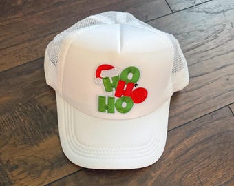 May include: White trucker hat with a mesh back and a curved brim. The front of the hat features the words "HO HO" in green and red, with a Santa hat graphic. A festive accessory for the holiday season.
