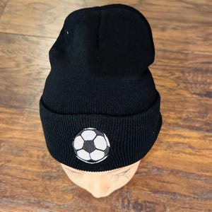 May include: Black knit beanie featuring a white and black soccer ball patch. The hat is made of a soft, ribbed material and is designed to fit snugly. A classic accessory for sports fans.