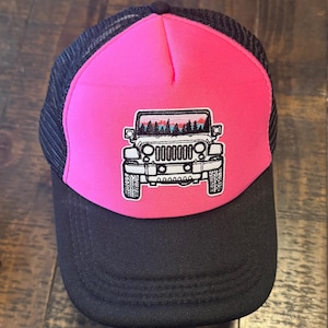 May include: A trucker hat with a black mesh back and a black brim. The front panel is bright pink with a white and black embroidered design of a Jeep with a forest scene.