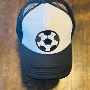 May include: A black and white trucker hat with a soccer ball graphic on the front. The hat features a black brim and mesh back, with a white front panel displaying a black and white soccer ball design.