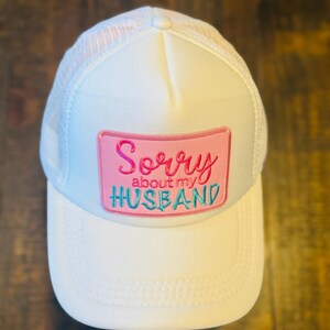 May include: White trucker hat with a mesh back and a curved brim. The front features a pink rectangular patch with the embroidered text "Sorry about my HUSBAND" in pink and teal lettering. The hat is made of a soft, breathable material.