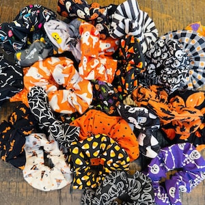 May include: A collection of Halloween-themed scrunchies in various patterns and colors. Designs include ghosts, spiders, candy corn, pumpkins, and skulls. The scrunchies are in shades of orange, black, white, and purple, perfect for festive hairstyles.