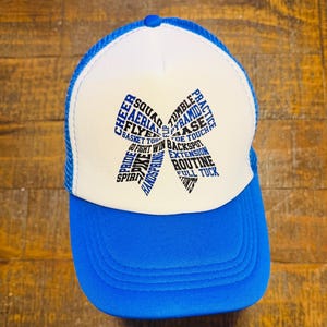 May include: Blue and white trucker hat with a curved brim. The hat features a white front panel with a blue bow-shaped design made of cheerleading terms like "CHEER", "SQUAD", and "TUMBLE". The mesh back is blue.