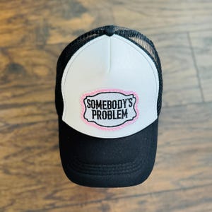 May include: Black and white trucker hat with a white front panel and black mesh back. The front panel features a pink-outlined patch with the text "SOMEBODY'S PROBLEM" in black.