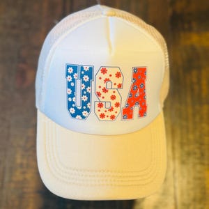 May include: A cream-colored trucker hat with a white mesh back and a curved brim. The front of the hat displays the word "USA" in red and blue, each letter filled with a floral pattern. The hat is designed with a classic trucker style.