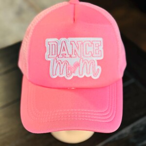 May include: A bright pink trucker hat with a mesh back and a curved brim. The front of the hat features the text "DANCE MOM" in white and pink lettering. The hat is displayed on a mannequin head.
