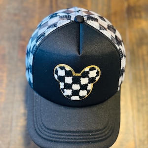May include: Black and gray trucker hat with a checkered pattern on the top and a black brim. The hat features a gold glitter Mickey Mouse head design with a checkered pattern.