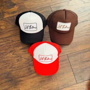 May include: Three trucker hats with the text "Let them" on a white patch. One hat is black with a white front, another is brown, and the third is red with a white front. The hats are on a wooden surface.