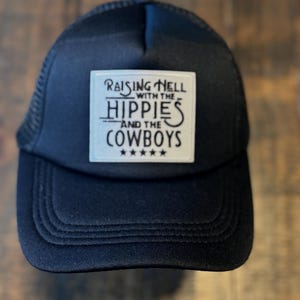 May include: Black trucker hat with a white rectangular patch. The patch features the text "Raising Hell with the Hippies and the Cowboys" in a stylized font, with five stars below the text. The hat has a mesh back.