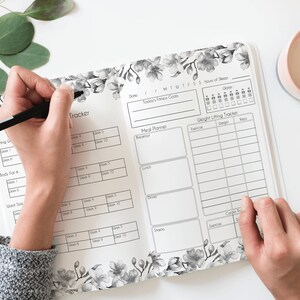 Pode incluir: Um planejador de design floral preto e branco com seções para acompanhar o sono, as refeições e o levantamento de peso. O planejador está aberto em uma página com um rastreador de levantamento de peso e um planejador de refeições.