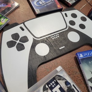 May include: A large, black and white wooden model of a PlayStation 5 controller. The controller is made of wood with a black and white paint scheme. The controller is sitting on a table with other video game cases.