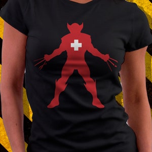 Puede incluir: Camiseta negra con una silueta roja de una figura de superhéroe con una cruz blanca en el pecho. La figura sostiene dos garras plateadas en cada mano.