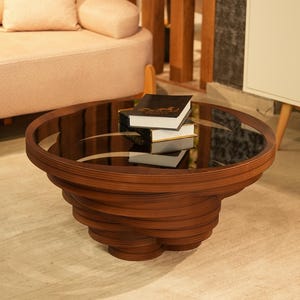 May include: A round coffee table with a dark brown wooden base composed of stacked, spiraling layers and a circular black glass top. Two stacked books with white pages and dark covers are placed on the center of the glass surface.
