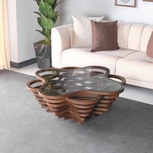 May include: A contemporary coffee table featuring a layered wooden base and a clear glass top. The table has an unusual, organic shape. The table is surrounded by a neutral-toned sofa and a potted plant.