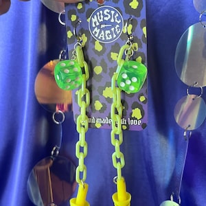 Puede incluir: Pendientes verde neón con dijes de dados y tapones para los oídos amarillos. Cuelgan de una tarjeta con el texto "MUSIC MAGIC" y un diseño de estampado de leopardo. Los pendientes están conectados por una cadena.