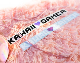 May include: Two pink and white pixelated stickers with the text "Kawaii Gamer" on a pink fuzzy surface. The stickers have a small purple pixelated heart in the middle.