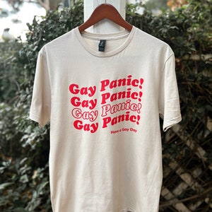 May include: A beige t-shirt with the phrase "Gay Panic!" repeated four times in red text. The text is in a retro font style. The shirt also has the text "Have a Gay Day" in smaller red text.