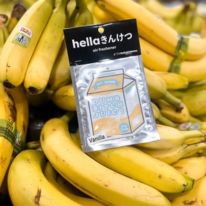 May include: A silver pouch with a white air freshener inside. The air freshener is shaped like a milk carton and has the text "99% DUMB DUMB BITCH JUICE" in blue and yellow letters. The pouch has the text "hellaきんけつ air freshener @hellakinketsu" in black letters. The air freshener is sitting on top of a pile of yellow bananas.