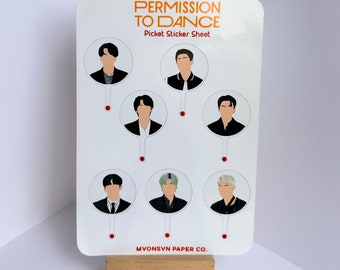 May include: A white sticker sheet with seven round stickers featuring illustrations of seven different people. The stickers are for a K-pop group called "Permission to Dance". The text "Picket Sticker Sheet" is printed in orange at the top of the sheet. The text "MVONSVN PAPER CO." is printed in red at the bottom of the sheet.