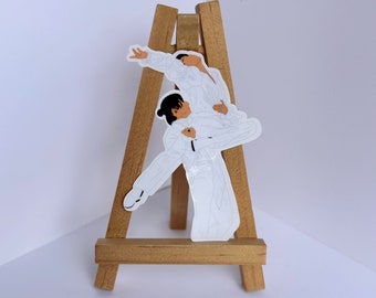May include: A white sticker on a wooden easel depicting two figures in a dance pose. The figures are outlined in black and are wearing white clothing.