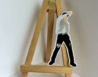 May include: A black and white illustration of a person wearing a white shirt, black pants, and a black belt. The illustration is printed on a white sticker and is placed on a small wooden easel.