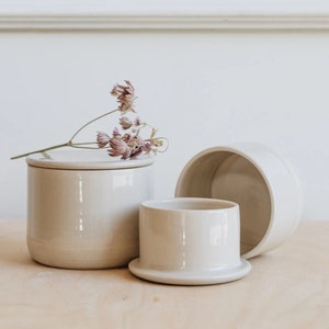 May include: A set of three ceramic butter dishes in a light beige colour. The dishes are stacked on top of each other, with the largest dish at the bottom and the smallest dish on top. The dishes have a smooth, rounded shape and a slightly textured surface.