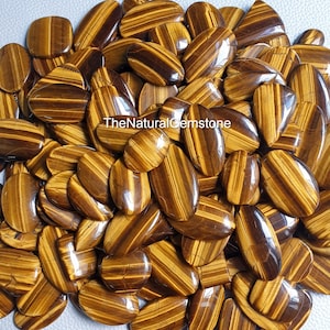 Op de afbeelding: Een grote stapel gepolijste tijgeroog-cabochons. De stenen zijn een mix van goudbruin en bruin met een chatoyante uitstraling. De stenen zijn ovaalvormig en hebben een gladde, gepolijste afwerking.