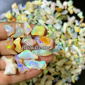 Peut inclure: Gros plan sur une main tenant un tas de petites pierres précieuses d'opale irisées. Les opales sont de différentes nuances de blanc, jaune et orange, et elles ont un effet chatoyant arc-en-ciel. Le texte "BulkGems" est visible sur l'image.