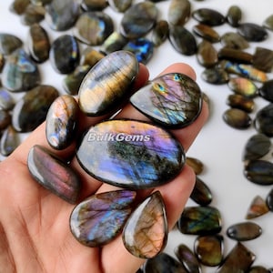 Puede incluir: Una colección de cabujones de labradorita, una piedra preciosa iridiscente, en varias formas y tamaños. Los cabujones son una mezcla de gris oscuro, morado y dorado con un efecto brillante. Los cabujones se muestran en una mano.