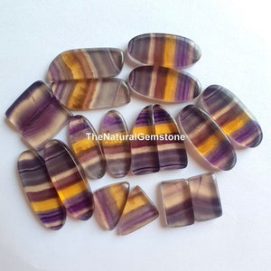 May include: A collection of 12 natural fluorite gemstones in various shapes and sizes. The stones are a mix of purple, yellow, and white with striations throughout. TheNaturalGemstone is printed on a white background.