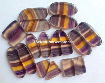 May include: A collection of 12 natural fluorite gemstones in various shapes and sizes. The stones are a mix of purple, yellow, and white with striations throughout. TheNaturalGemstone is printed on a white background.