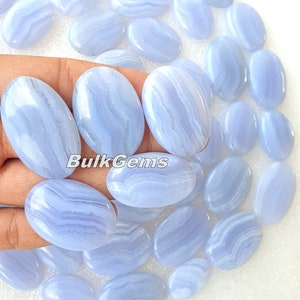 Op de afbeelding: Een verzameling blauwe agaat cabochons, ovaalvormig, met witte en blauwe banden. De cabochons zijn glad en gepolijst. De afbeelding toont een hand die verschillende cabochons vasthoudt. De tekst "BulkGems" is zichtbaar in de afbeelding.