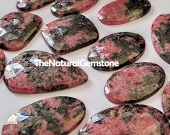 Puede incluir: Una colección de 12 cabujones de piedras preciosas rosas y negras. Las piedras están talladas en forma ovalada facetada y tienen una textura natural y rugosa. TheNaturalGemstone está impreso en el fondo blanco.
