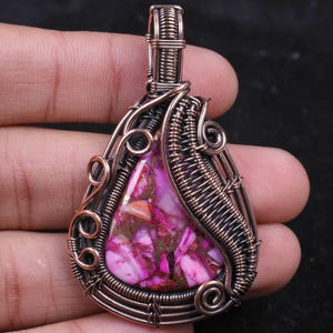 Peut inclure: Un pendentif artisanal avec une pierre en forme de goutte d'eau aux tons roses, violets et bruns. La pierre est sertie dans un fil de cuivre complexe, créant un bijou unique.