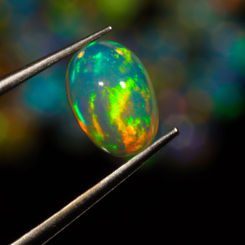 May include: A close-up of a green and orange opal gemstone held by tweezers. The opal is oval-shaped and has a translucent, iridescent appearance.
