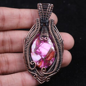 Peut inclure: Un pendentif composé d'une pierre ovale aux motifs marbrés rose vif, blanc et brun rougeâtre. La pierre est enchâssée dans une armature complexe en fil de cuivre oxydé foncé, formant des volutes et des boucles décoratives autour du centre.