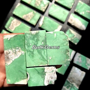 May include: A collection of green and white variscite cabochons, cut into rectangular shapes. The stones have a natural, earthy look with a mix of green and white colors. The cabochons are arranged in a grid pattern, with the text "BulkGems" visible in the center.