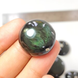 K&ouml;nnte beinhalten: Eine runde, polierte schwarze Obsidiankugel mit gr&uuml;nem und schimmerndem Schimmer. Die Kugel hat einen Durchmesser von etwa 5 cm.