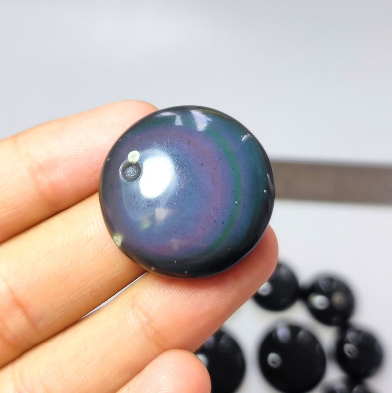 K&ouml;nnte beinhalten: Ein runder, irisierender Obsidian-Stein in dunkelblau, violett und gr&uuml;n. Der Stein hat einen wei&szlig;en Punkt in der Mitte.