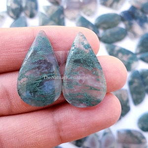 Puede incluir: Dos cabujones de ágata verde musgo en forma de lágrima con un aspecto translúcido y nublado. Las ágatas tienen un patrón natural y orgánico de verde y blanco.