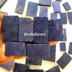 Puede incluir: Una colección de virutas de lapislázuli azul oscuro, algunas de las cuales se sostienen en una mano. Las virutas son rectangulares y tienen una superficie rugosa y texturizada. El texto "BulkGems" es visible en la imagen.