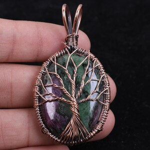 May include: An oval pendant featuring a polished green and purple stone wrapped in copper wire. The wire is intricately shaped into a tree design with branches extending across the stone surface, secured by a copper wire frame and a top loop.
