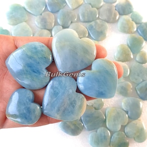 Puede incluir: Una colección de gemas en forma de corazón de aguamarina. Las piedras pulidas varían en tonos de azul claro y se sostienen en una mano. Las gemas tienen vetas naturales y se muestran sobre un fondo blanco. El texto "Bulk Gems" es visible.