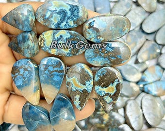 May include: A collection of blue, brown, and white gemstone cabochons. The cabochons are in various shapes, including oval and teardrop. The cabochons are arranged in a pile on a hand.
